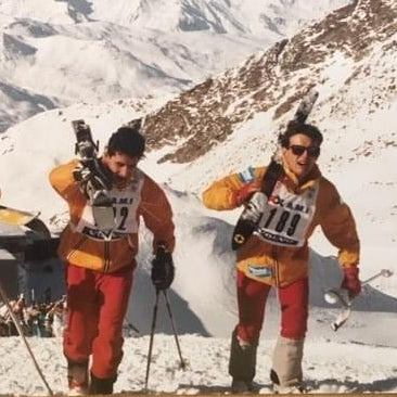 Three skiers in orange and yellow outfits with ski equipment on a snowy mountain background

